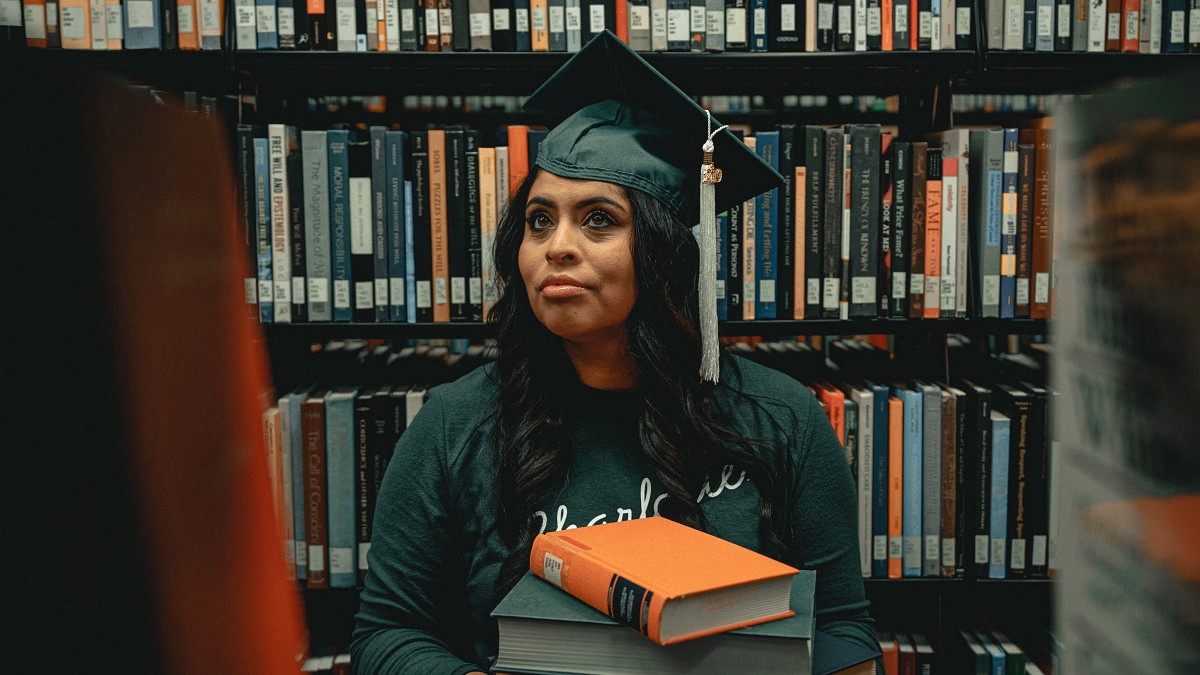 student wearing green cap in a library holding multiple books on her hand