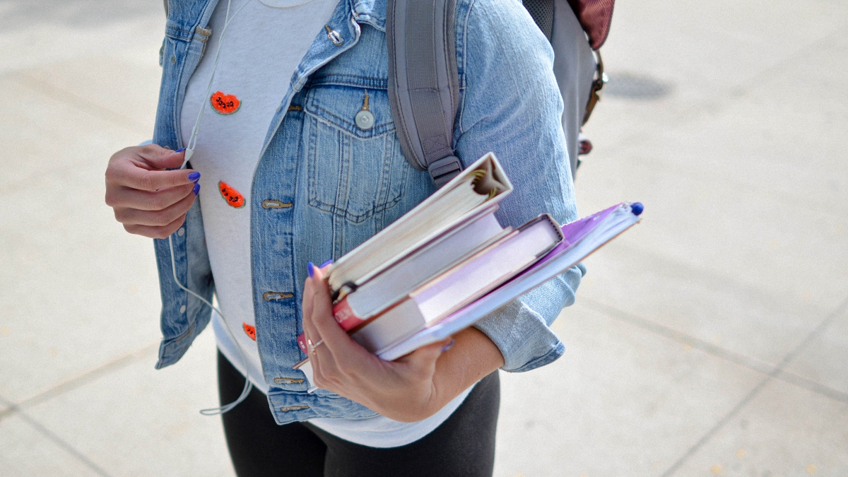 A high school student wearing blue jean jacket holding books on hand